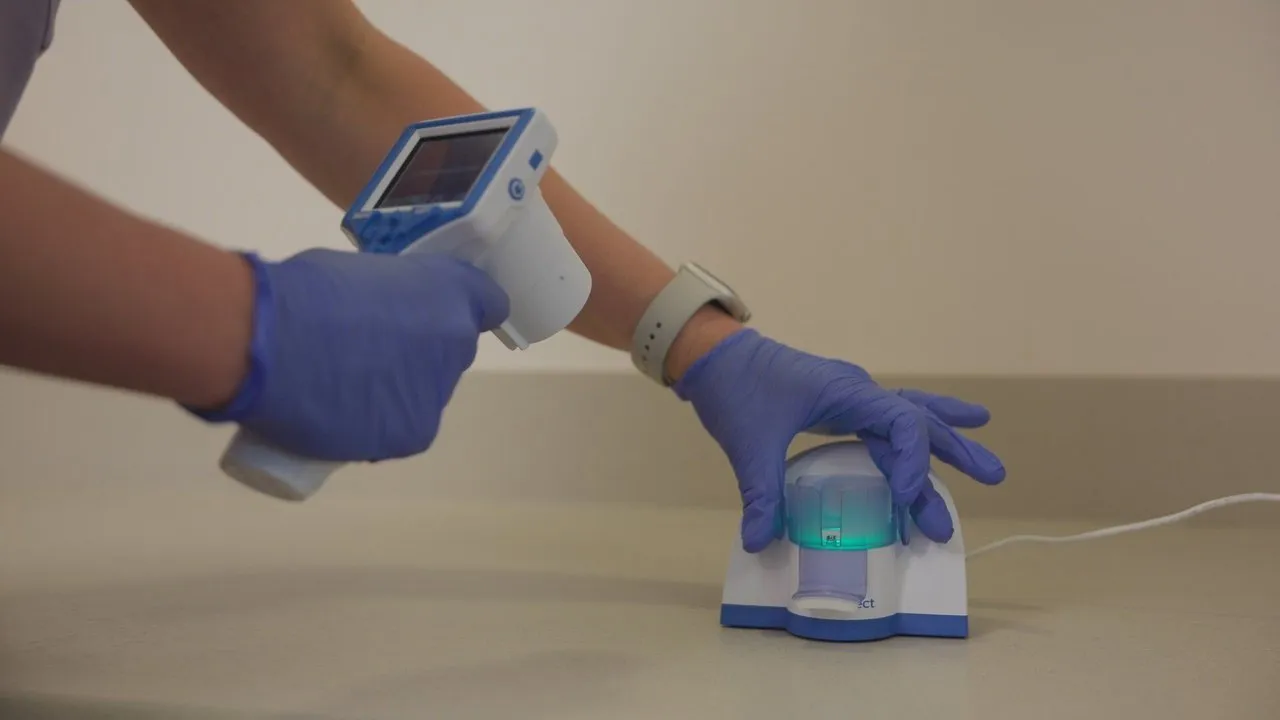 Gloved hands use a handheld scanner beside a small connected medical device on a countertop.