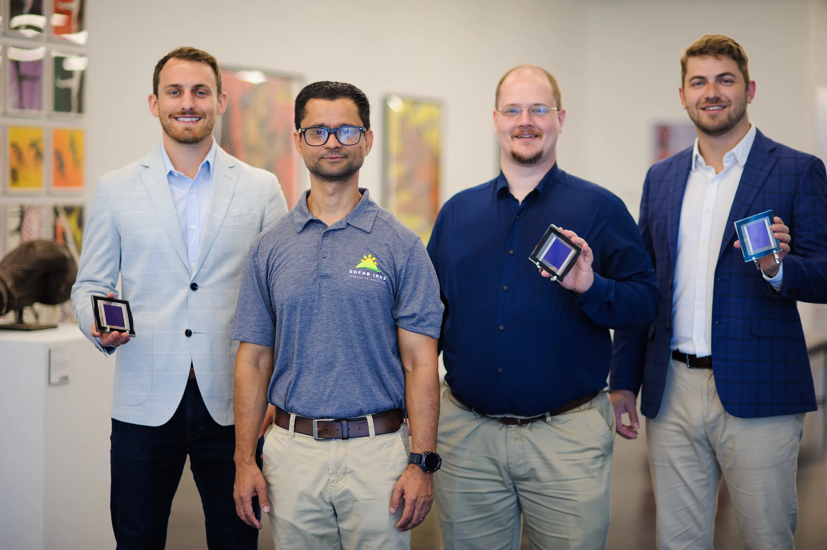 Four men stand in an office, three holding small square electronic devices.