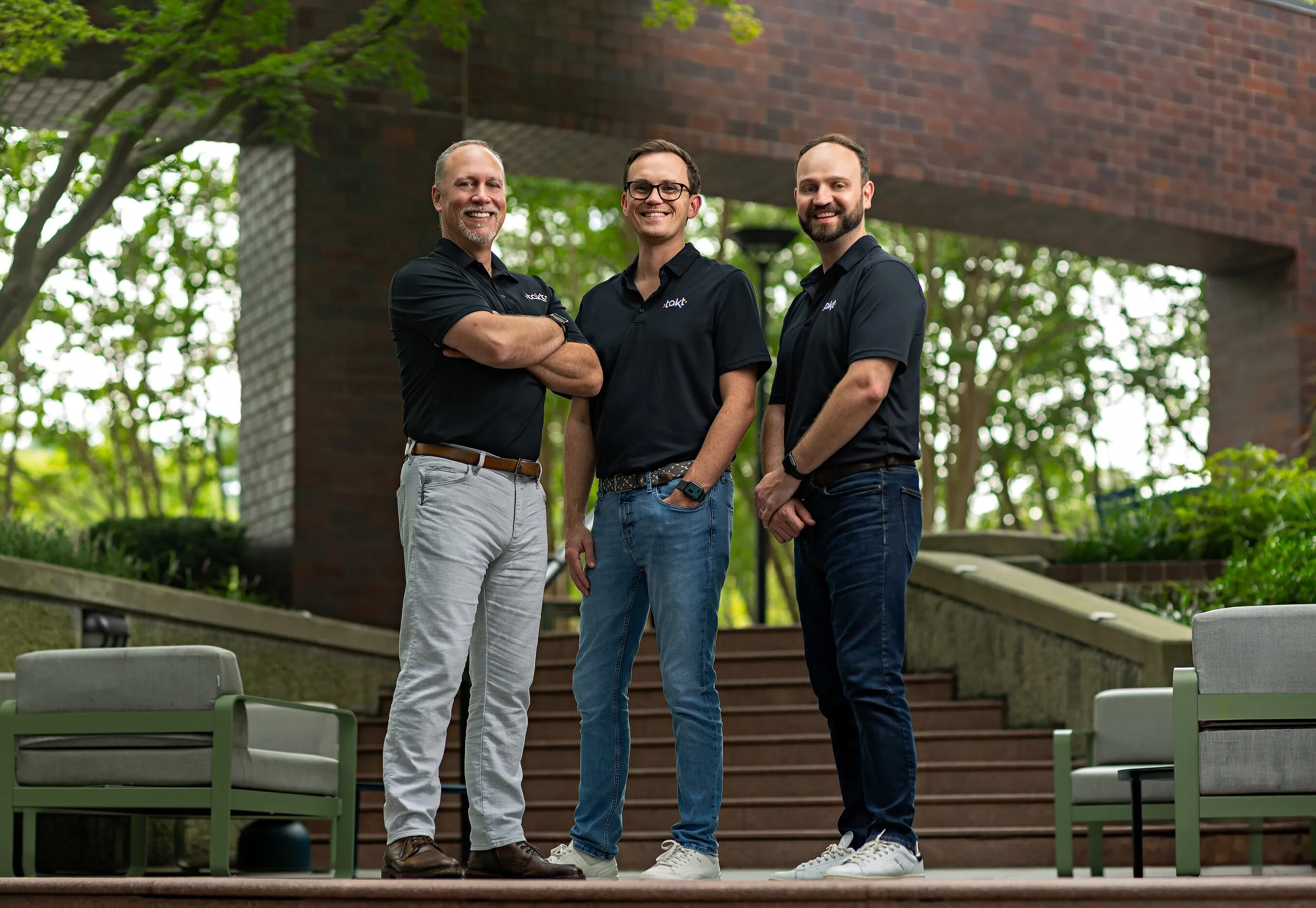 Three men in black Takt polo shirts standing outdoors by brick walls and chairs.