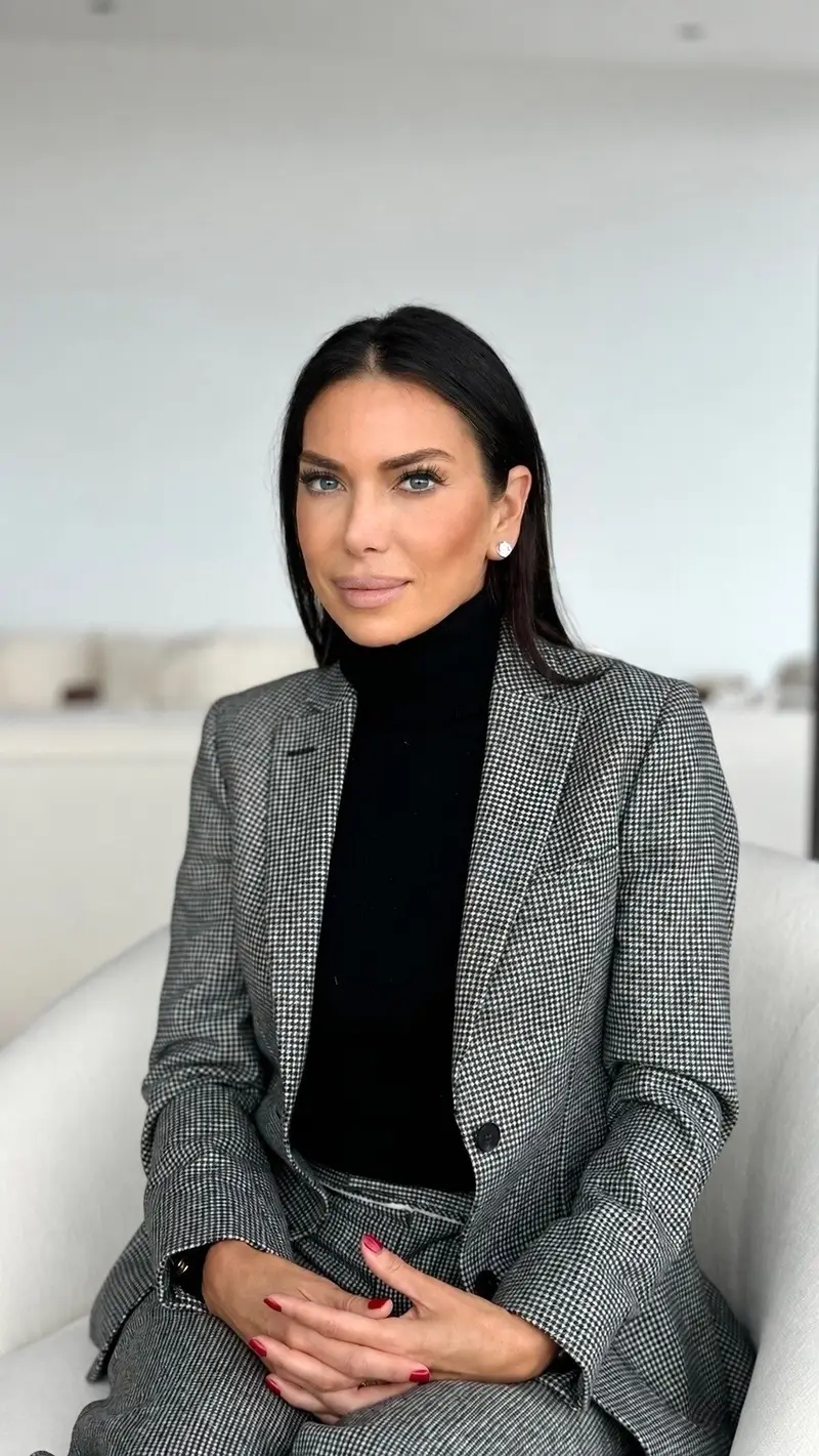 Tanya Zuckerbrot in a black turtleneck and checked blazer, seated on a white chair.