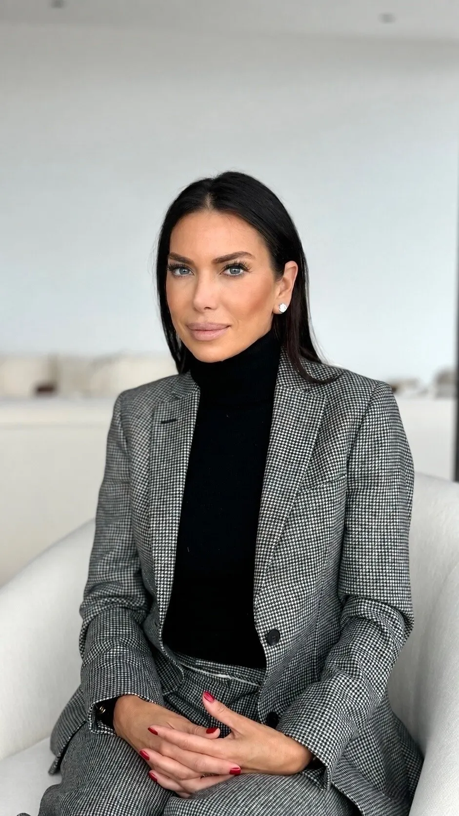 Tanya Zuckerbrot in a black turtleneck and checked blazer, seated on a white chair.