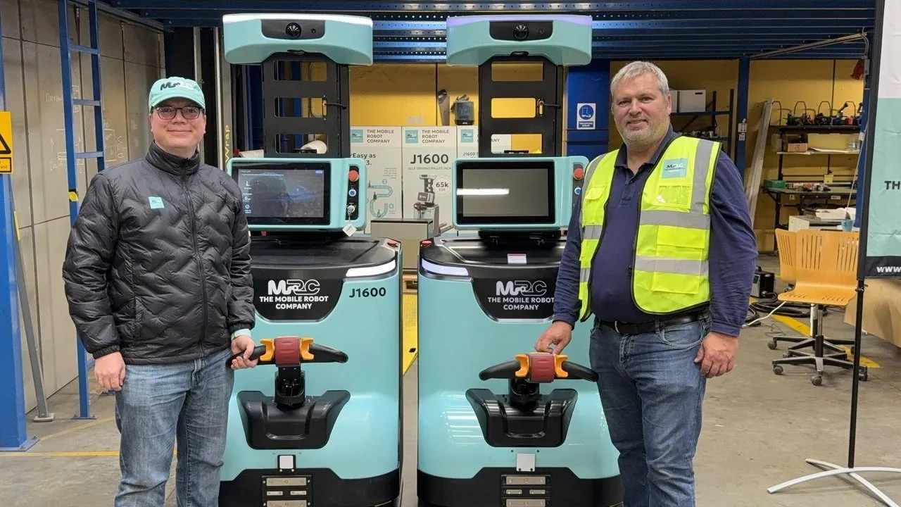 Two people stand beside two Mobile Robot Company J1600 warehouse robots.