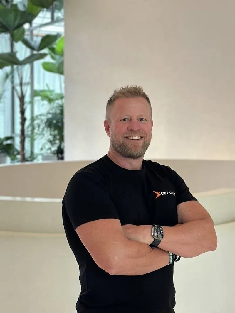 Troy Adams wearing a black Crossware T-shirt, standing with arms crossed indoors beside plants.