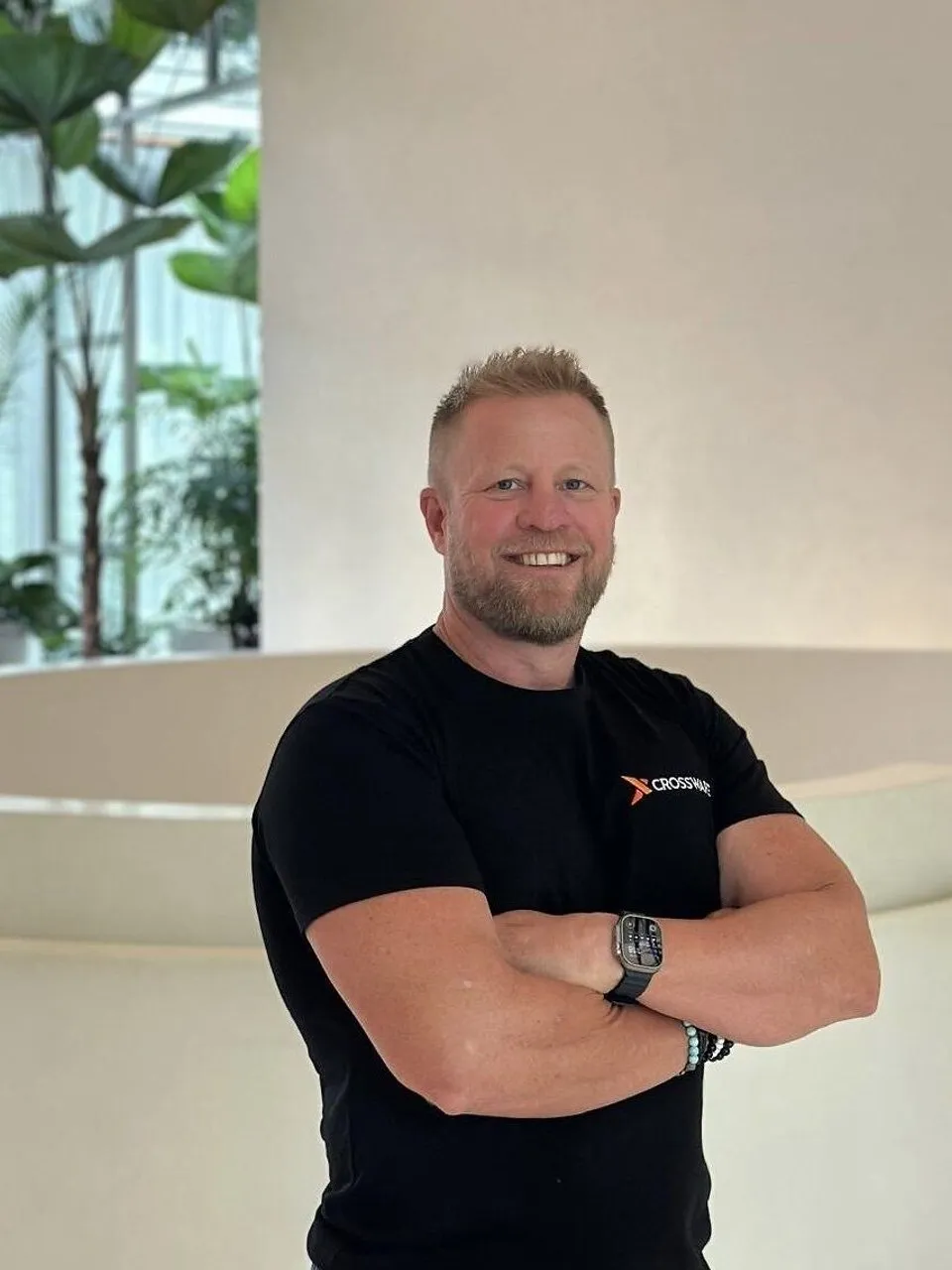 Troy Adams wearing a black Crossware T-shirt, standing with arms crossed indoors beside plants.