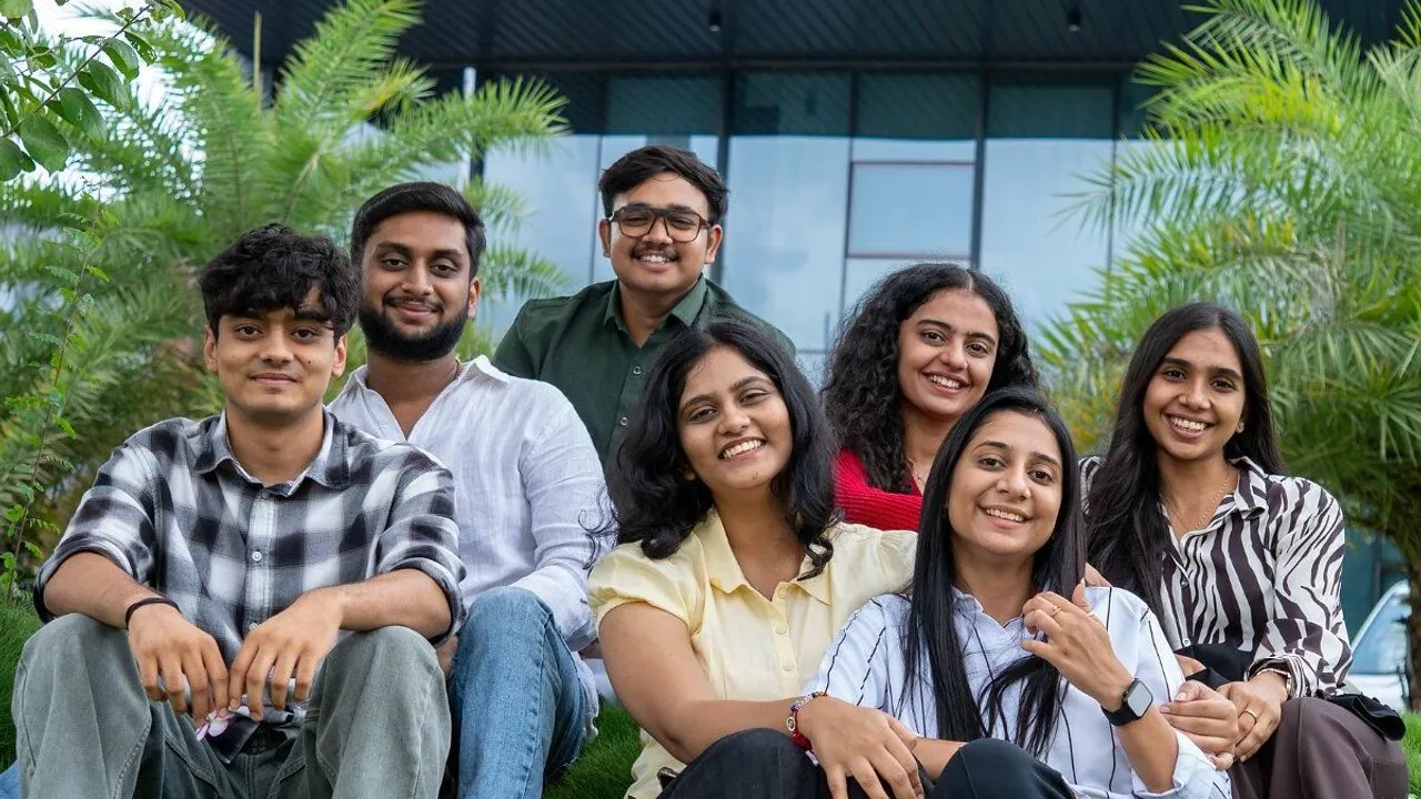 Seven young adults smiling together outdoors amid palm trees.