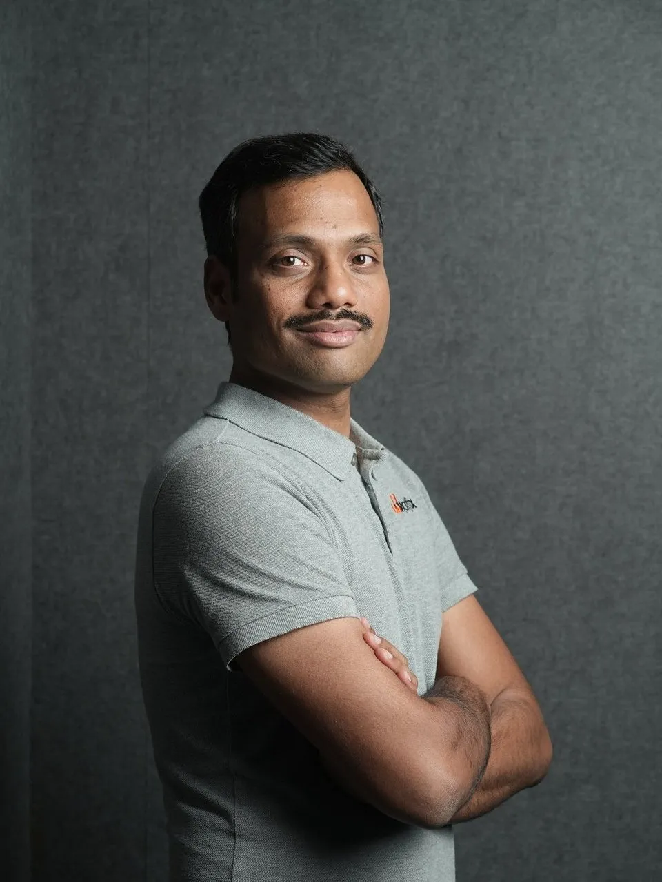 Vara Kumar wearing a gray polo shirt with arms crossed against a gray backdrop.