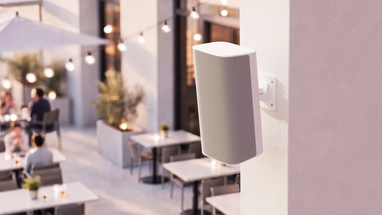 White wall-mounted speaker overlooking an outdoor restaurant patio with tables and string lights.
