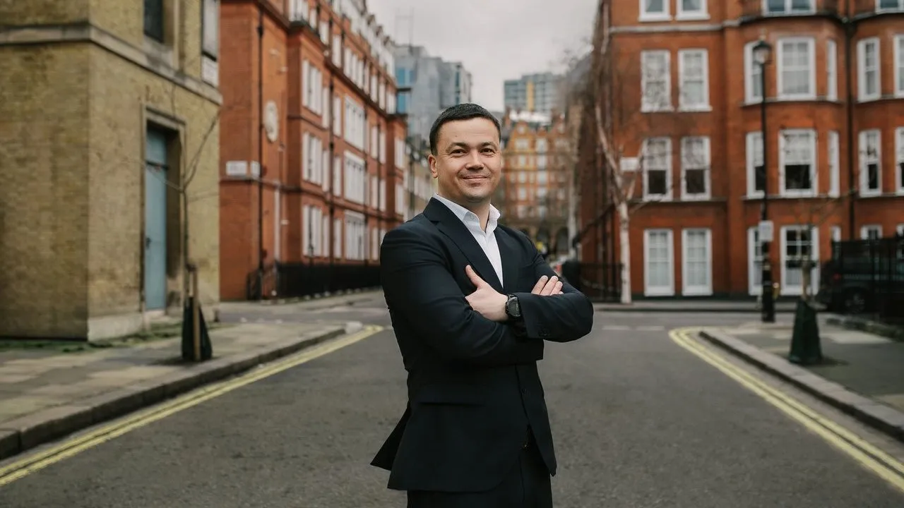Tim Khamzin stands with arms crossed on a city street lined with red-brick buildings.