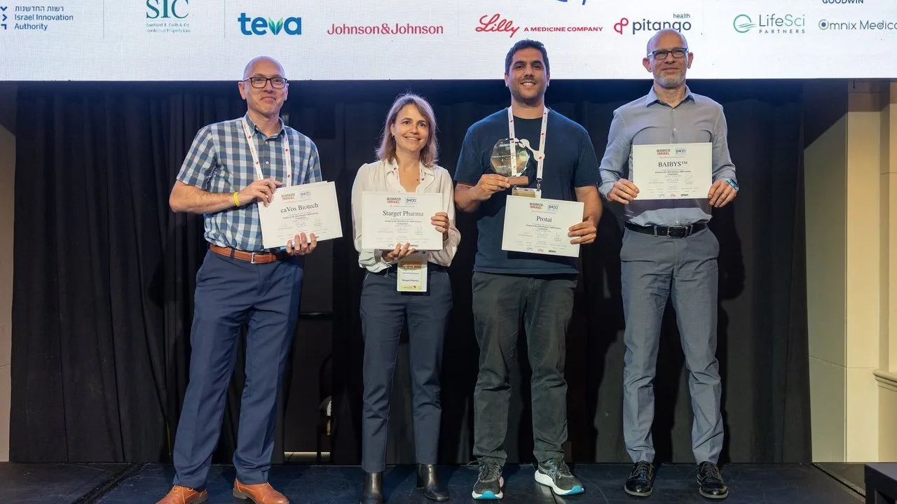 Four award winners holding certificates and a trophy on a stage beneath sponsor logos.