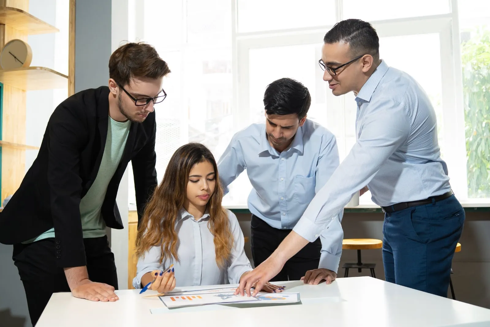 A young woman at a table with a printed chart while three men lean over it, one pointing to the page