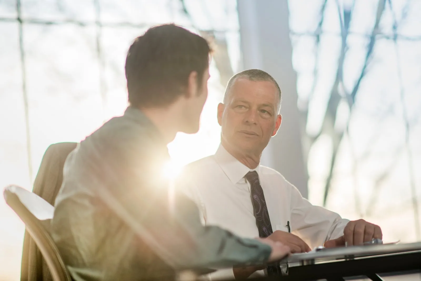 An older man in a white shirt and tie listens to a younger man across a table in a bright office