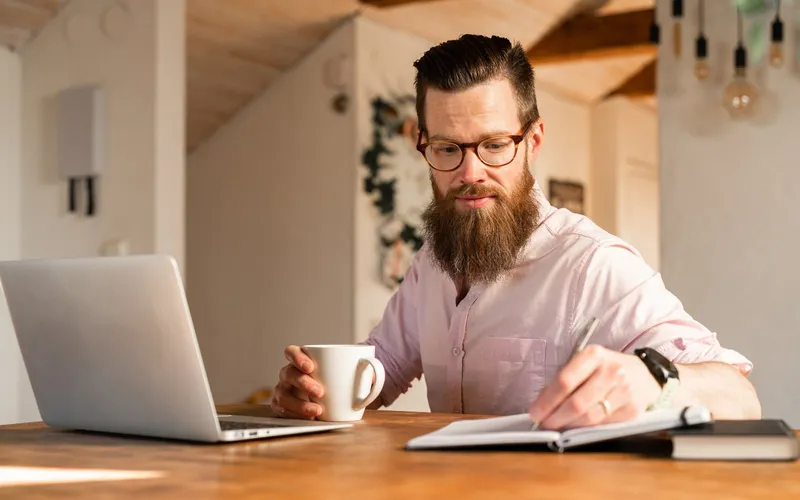 A man with coffee and a notebook at his laptop