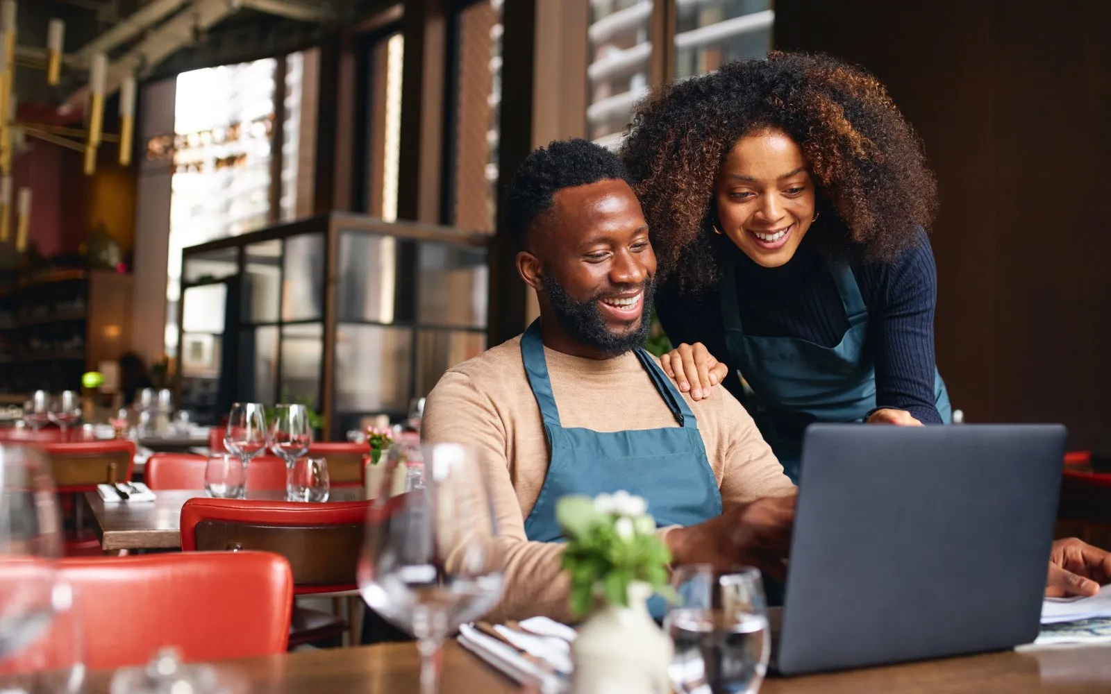 Two café owners in aprons smiling at a laptop in their restaurant