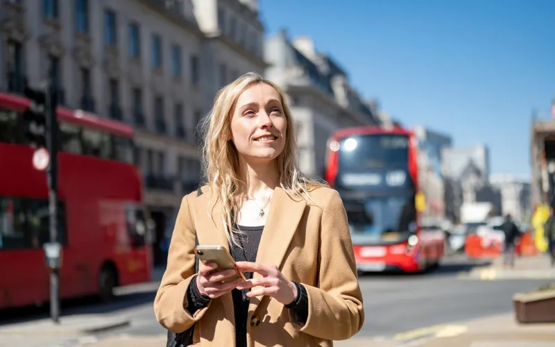 A woman with her phone on a London street, red buses behind
