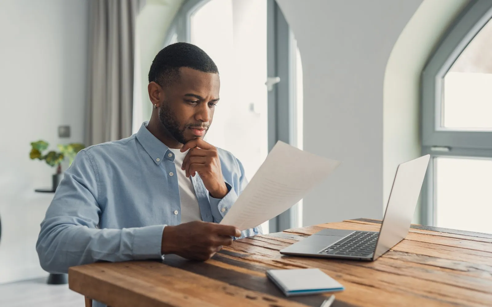 A man reading a document at a wooden desk beside his laptop