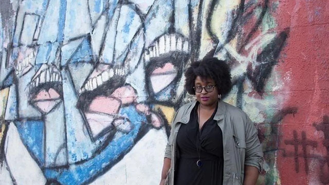 A woman stands in front of a colorful mural of abstract faces with gritted teeth.