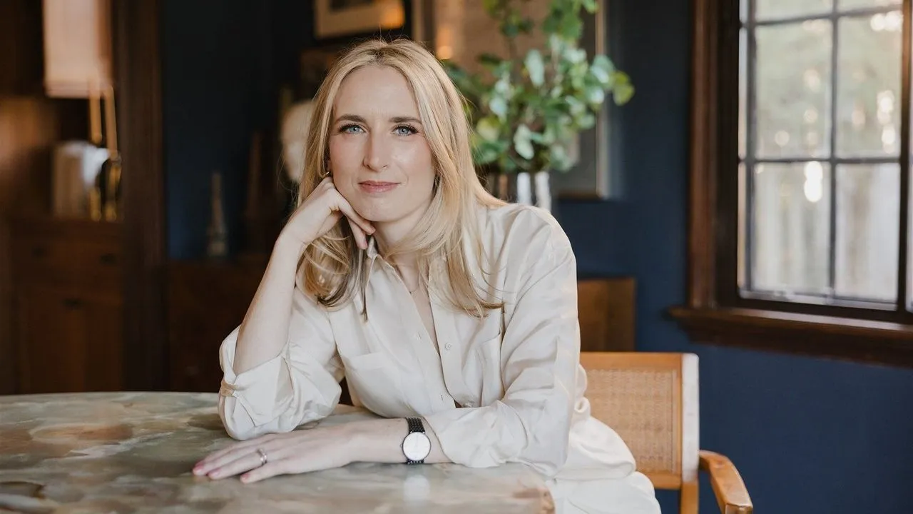 Blonde woman in a white shirt seated at a marble table, resting her chin on her hand.