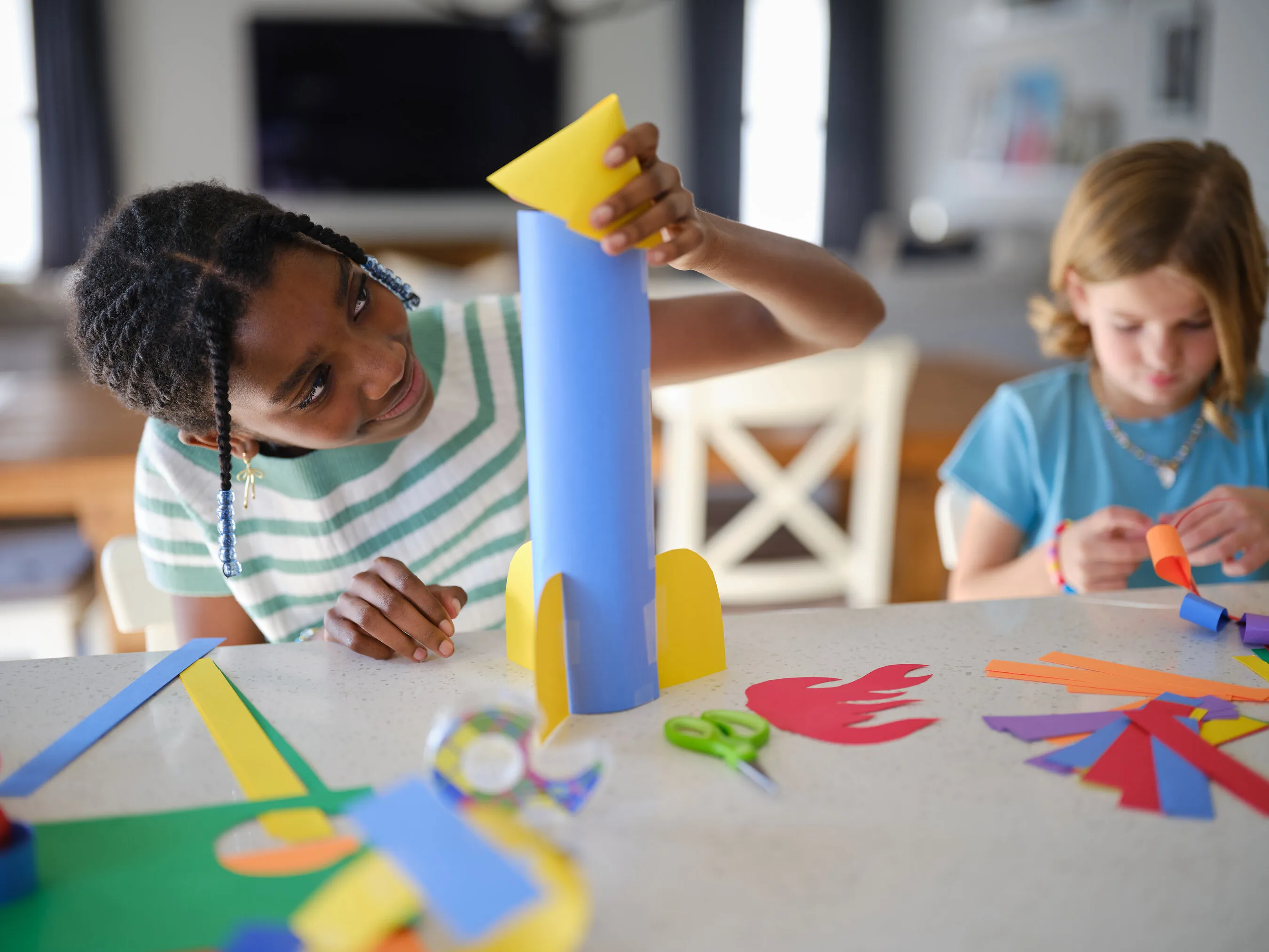 Two children make colorful paper crafts at a table, including a blue and yellow paper rocket.